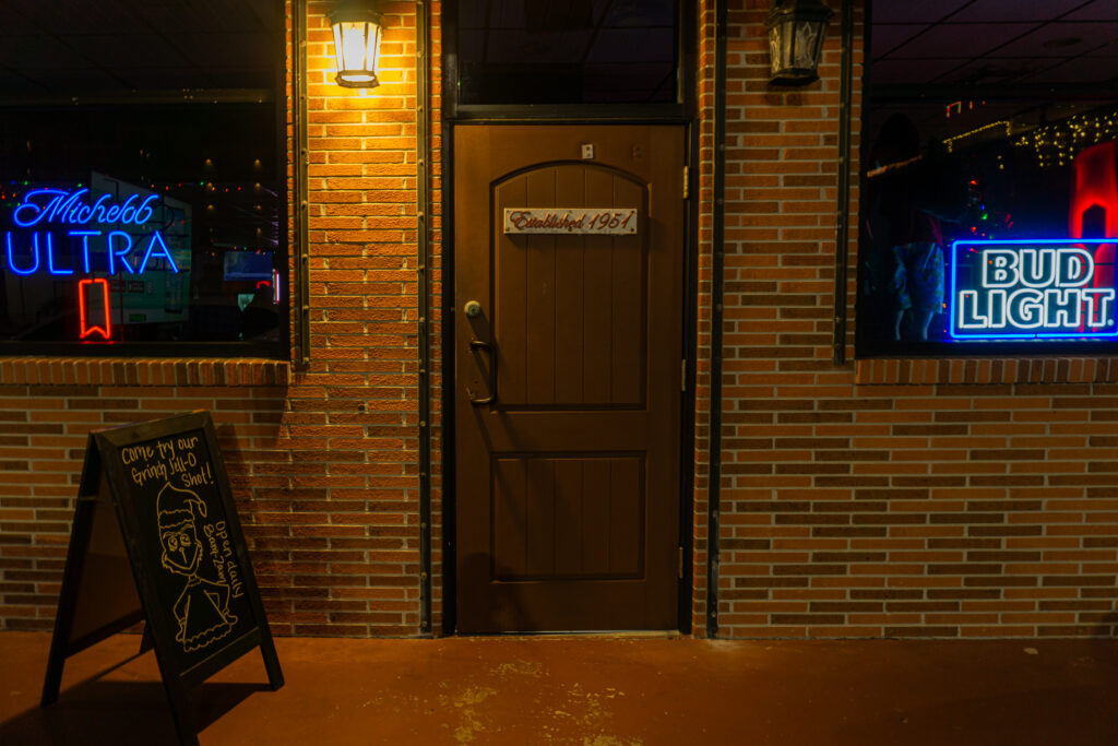 night shot of metz 1 front door. neon blue signs in windows. wooden door that says "established 1951".